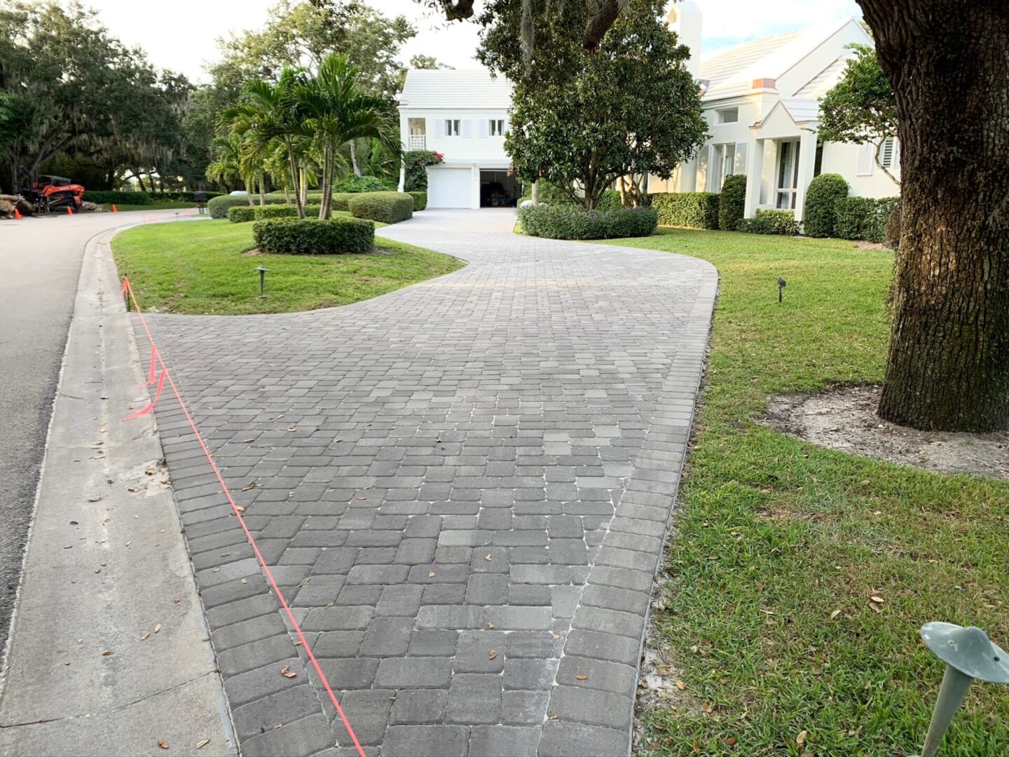 A wide brick-paved driveway leads to a garage and is bordered by a manicured lawn and shrubbery. Red tape is strung along one side, possibly indicating construction or maintenance work.