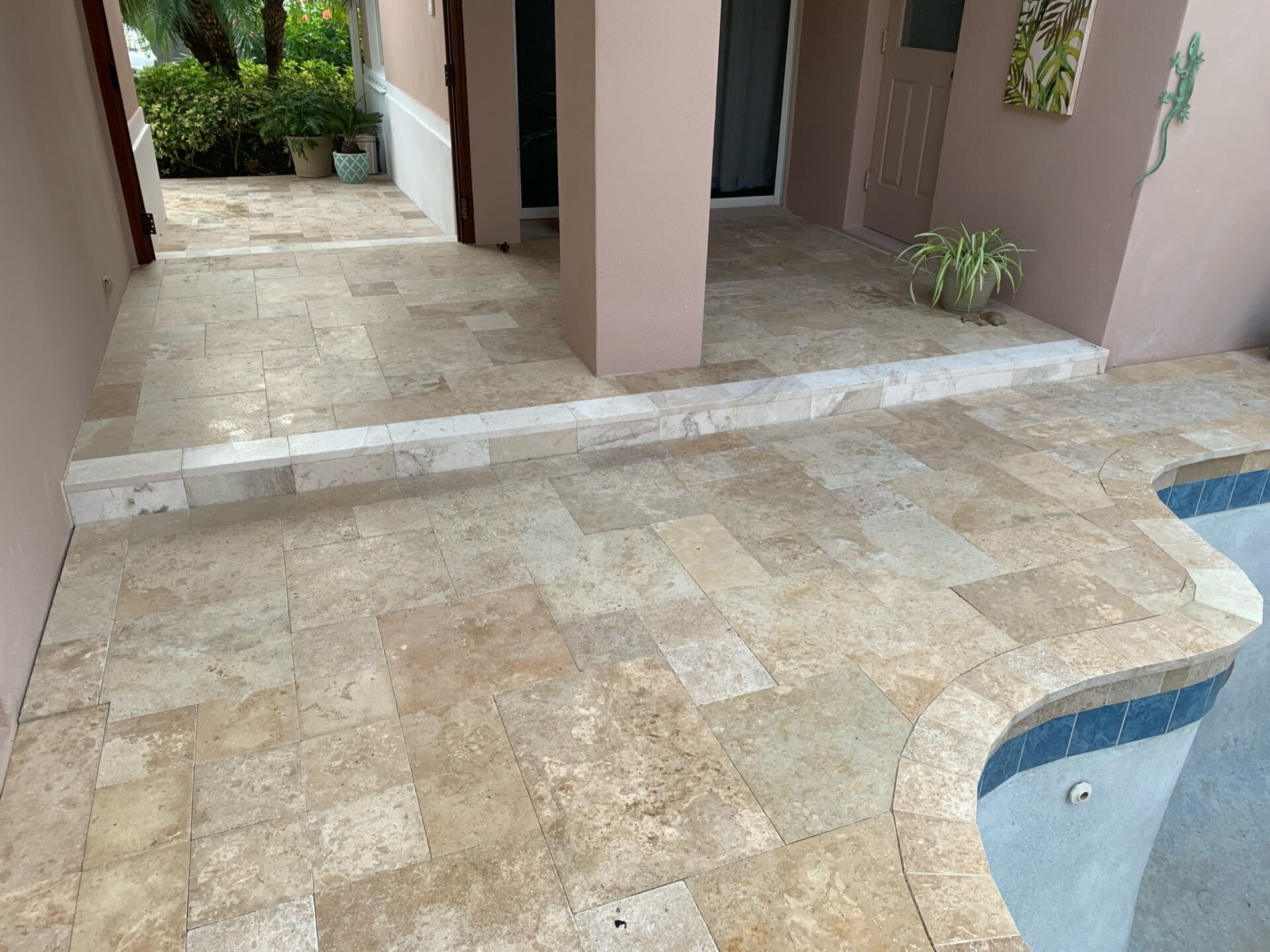 Outdoor patio area with beige and tan stone tile flooring near a pool. Features steps, a door, potted plants, and a decorative lizard on the wall.