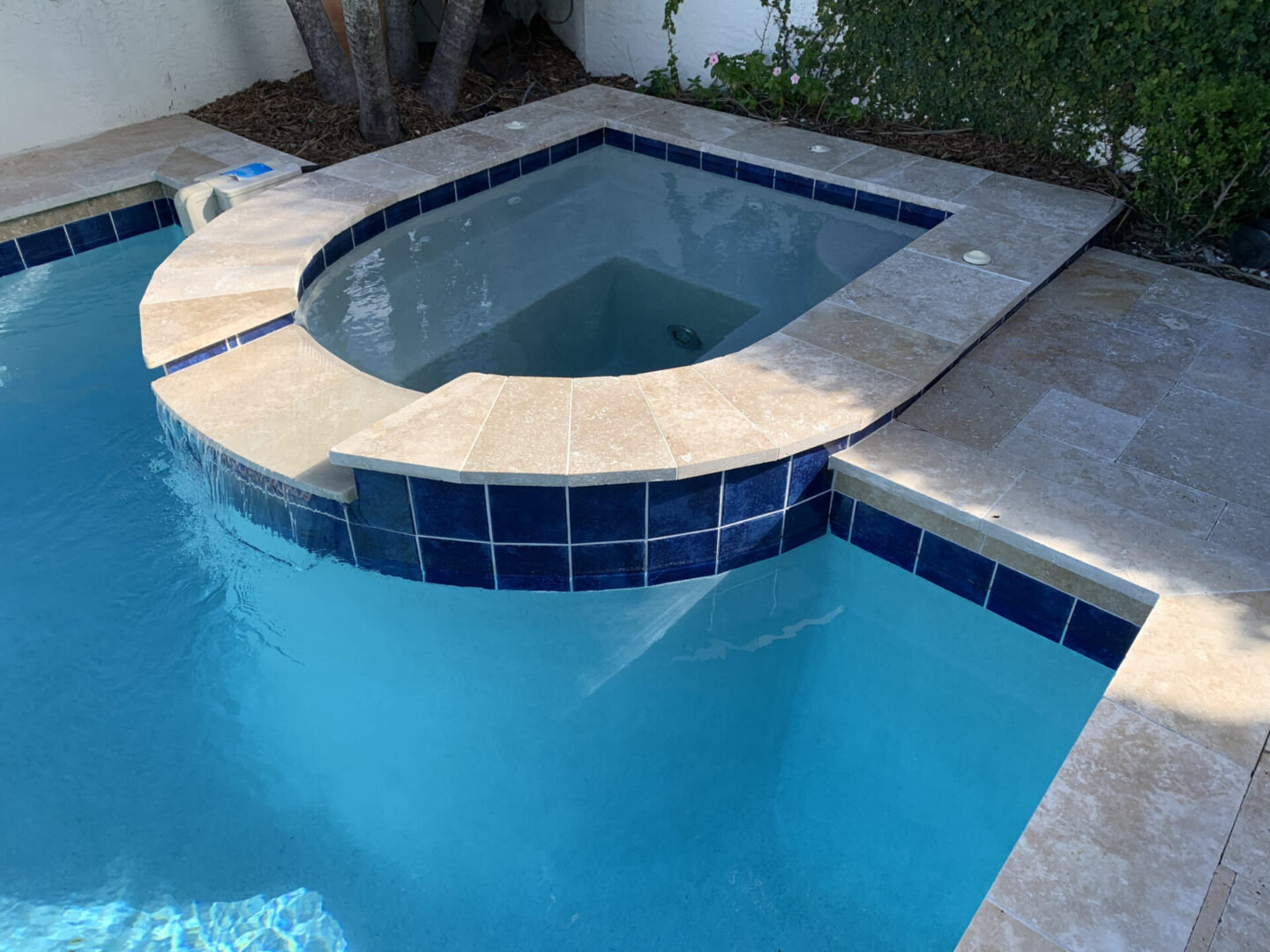 A small, elevated spa with blue tile borders connects to a larger swimming pool. Both have clear water and are surrounded by beige stone decking.