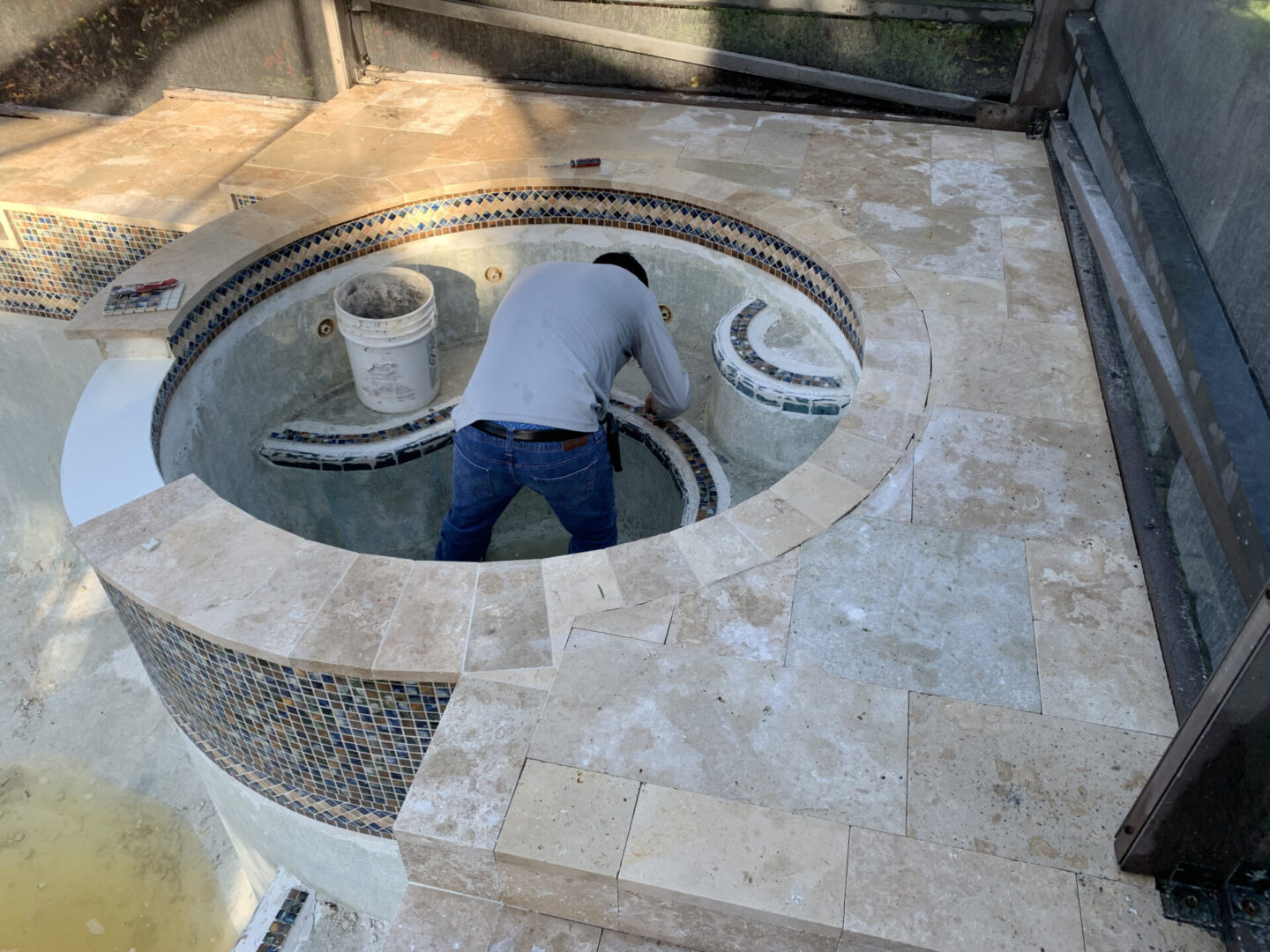 A person wearing a gray shirt and jeans works inside an empty, circular hot tub. The surrounding area features tiled edges and a bucket situated inside the tub.
