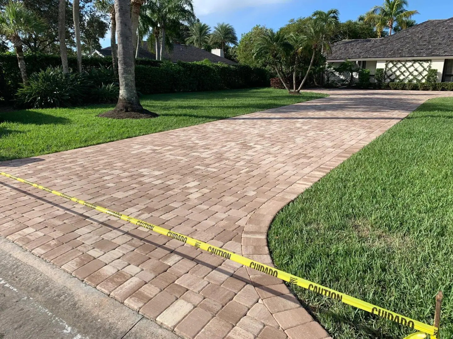 Brick driveway cordoned off with yellow caution tape, leading to a house surrounded by greenery, palm trees, and a cloudy sky.