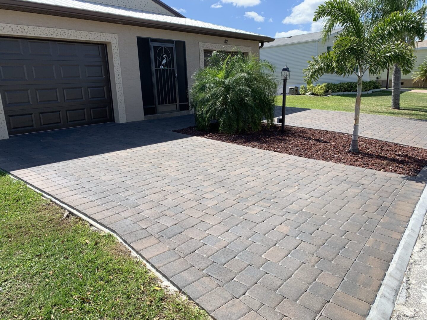 A home with a paver driveway and a single-car garage. A palm tree and a small lamp post are in a mulched area next to the driveway.
