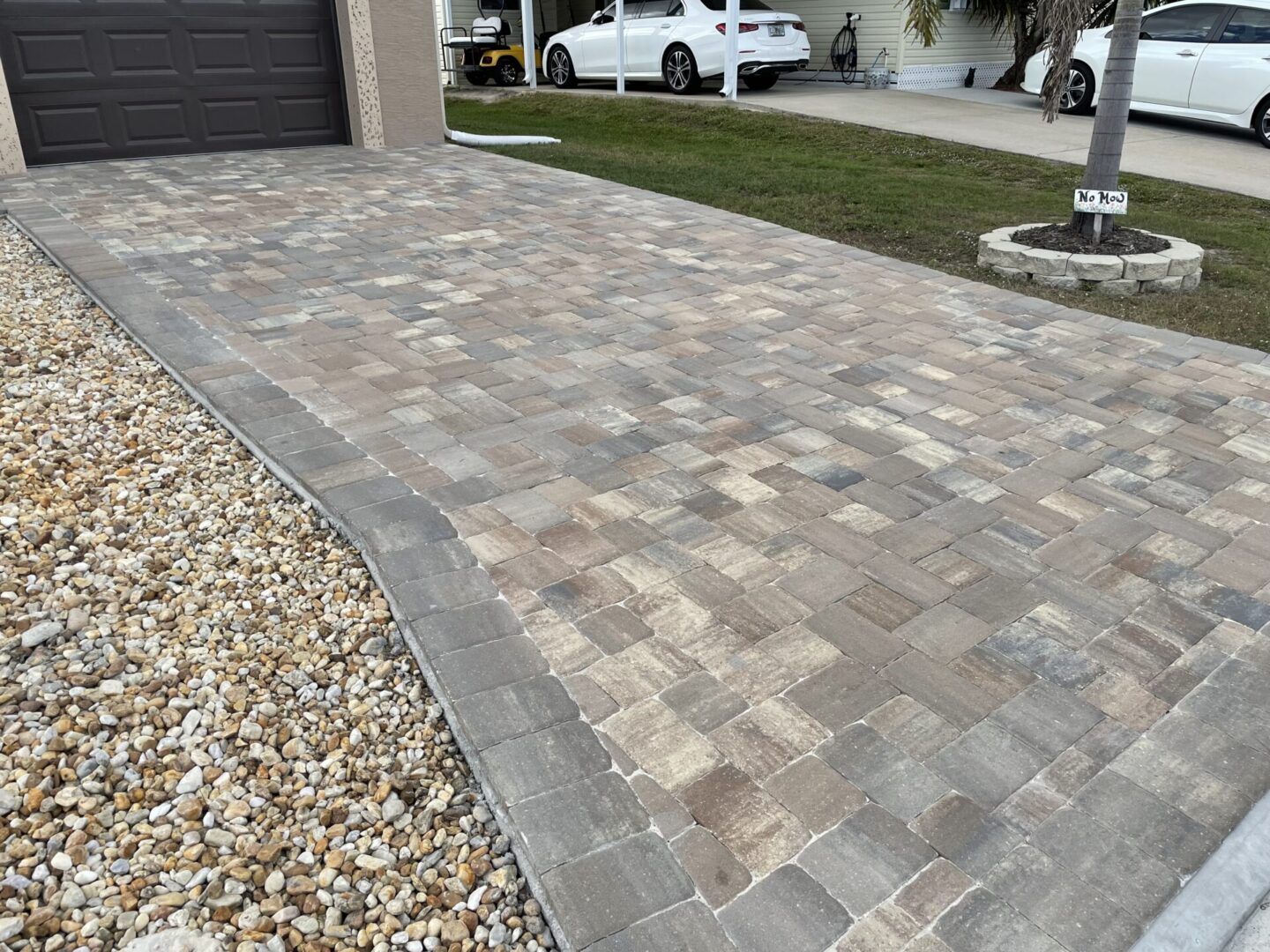 A driveway paved with interlocking stone tiles is bordered by pebbles on the left and a small patch of grass on the right, leading up to a closed garage door. Two cars are parked near a house in the background.
