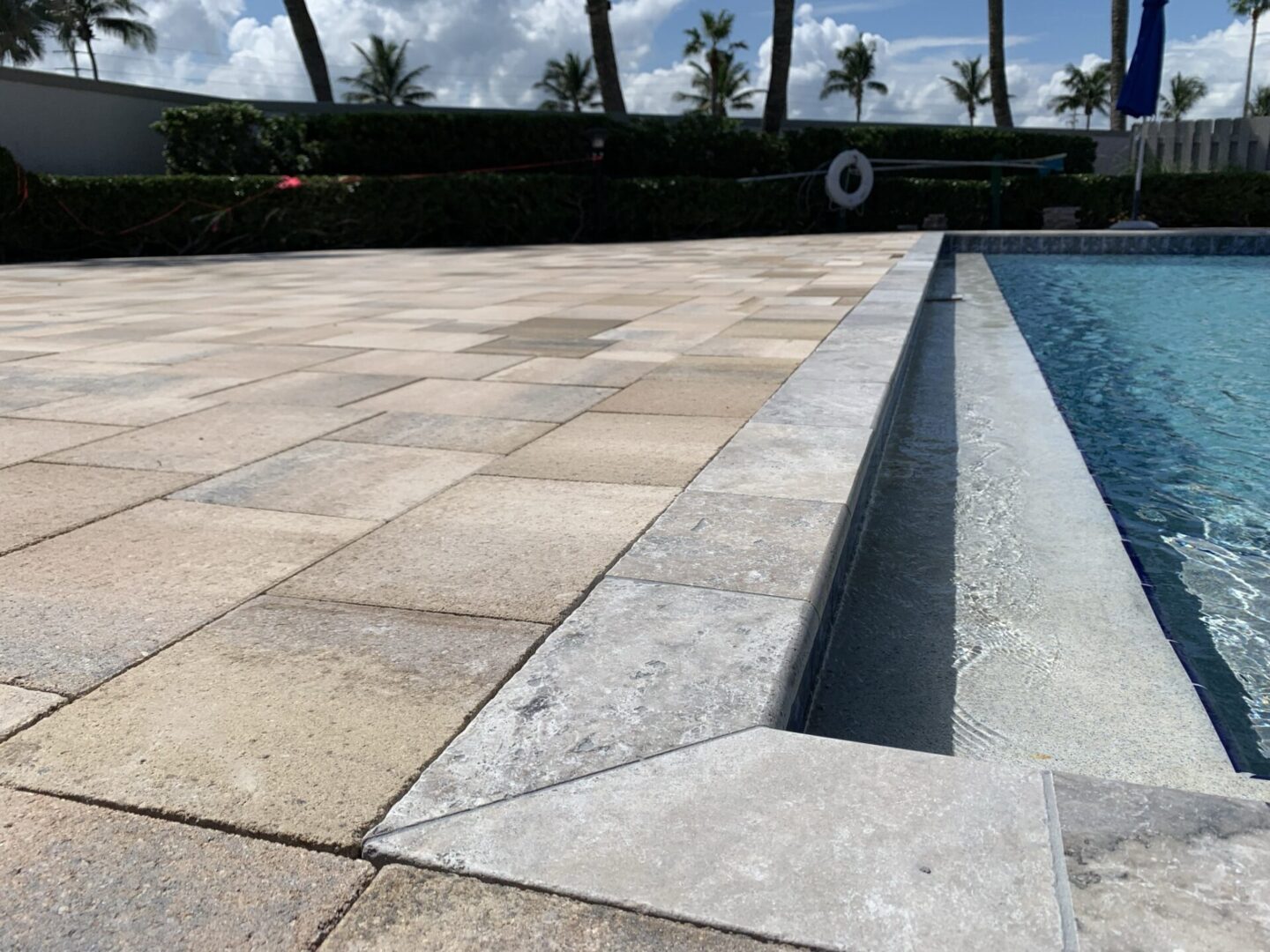 Close-up of a poolside area with paved stone tiles next to the edge of a swimming pool, surrounded by greenery, palm trees, and a cloudy sky in the background.