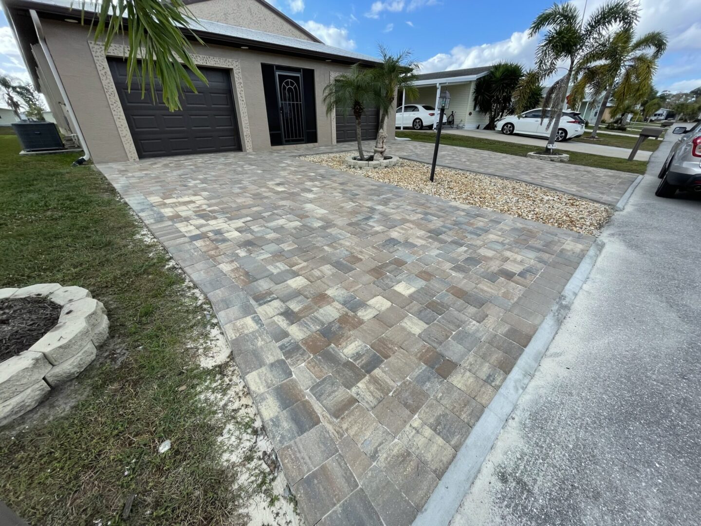 A paved driveway with a mix of beige and gray bricks leading up to a garage door. There are palm trees and a neatly landscaped garden area adjacent to the driveway.
