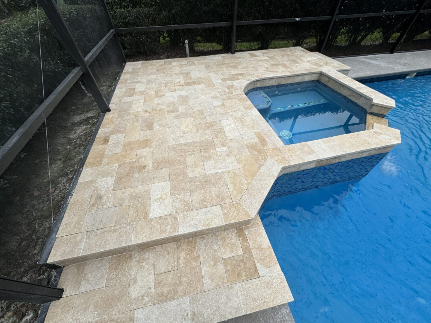 A swimming pool with an attached hot tub, surrounded by beige stone tiles and enclosed by a black mesh fence.