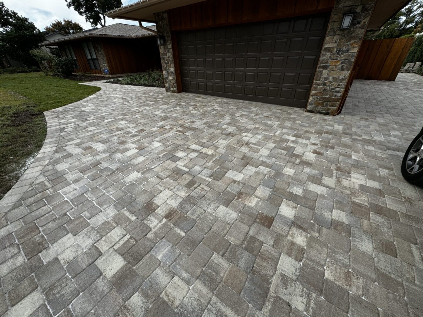 A driveway with a light brown, interlocking brick paver design leads to a house with a double garage door.