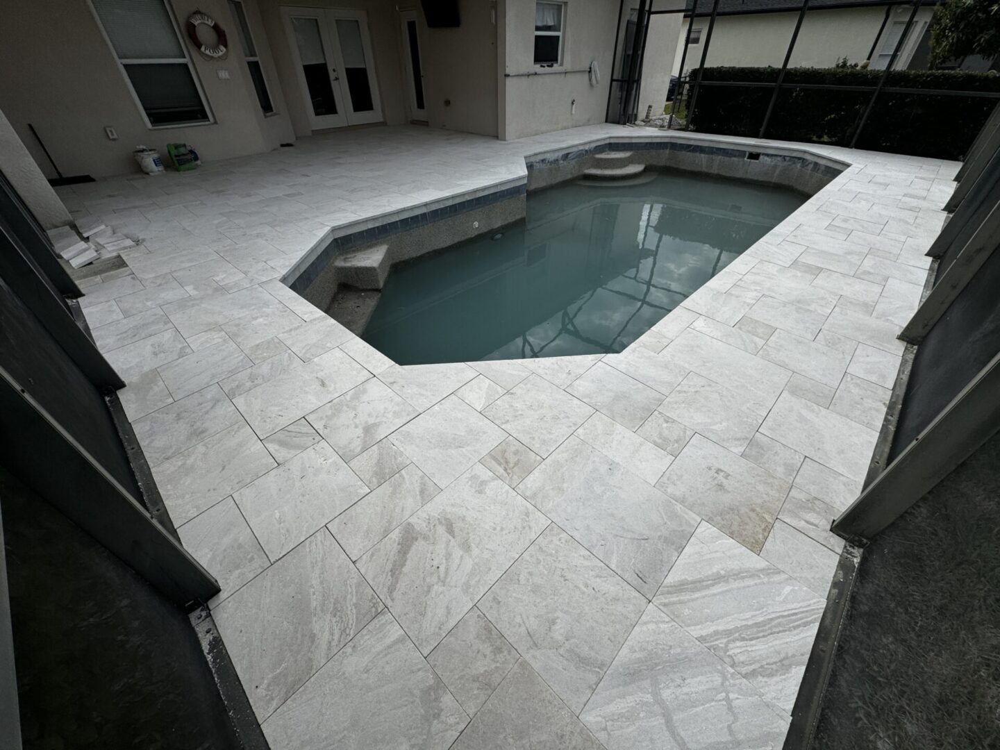A backyard with a partially filled, small, rectangular pool surrounded by new grey tile flooring. The area is enclosed by a screen.
