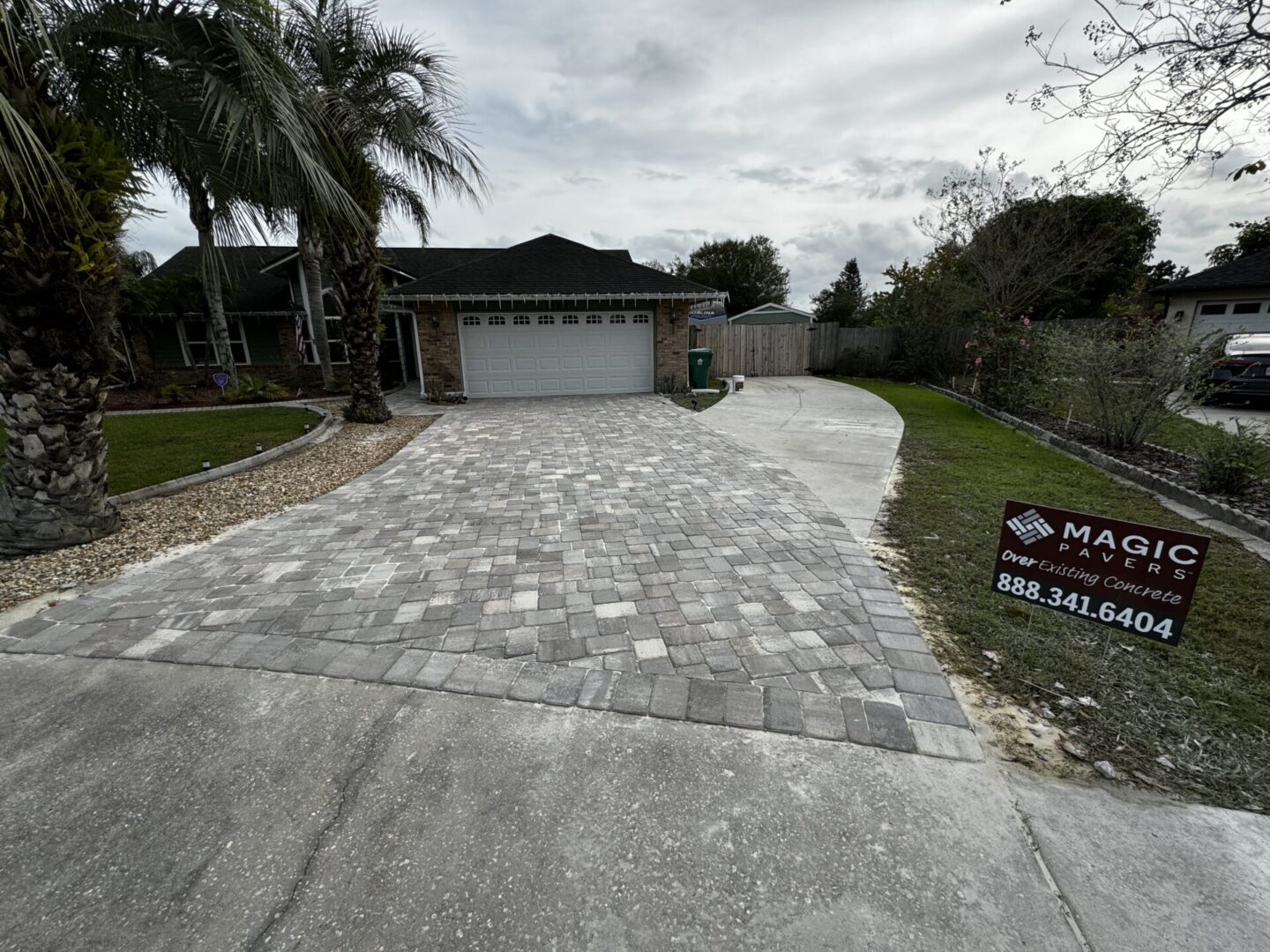 A house with a newly paved stone driveway and garage is shown. A sign on the lawn reads, 