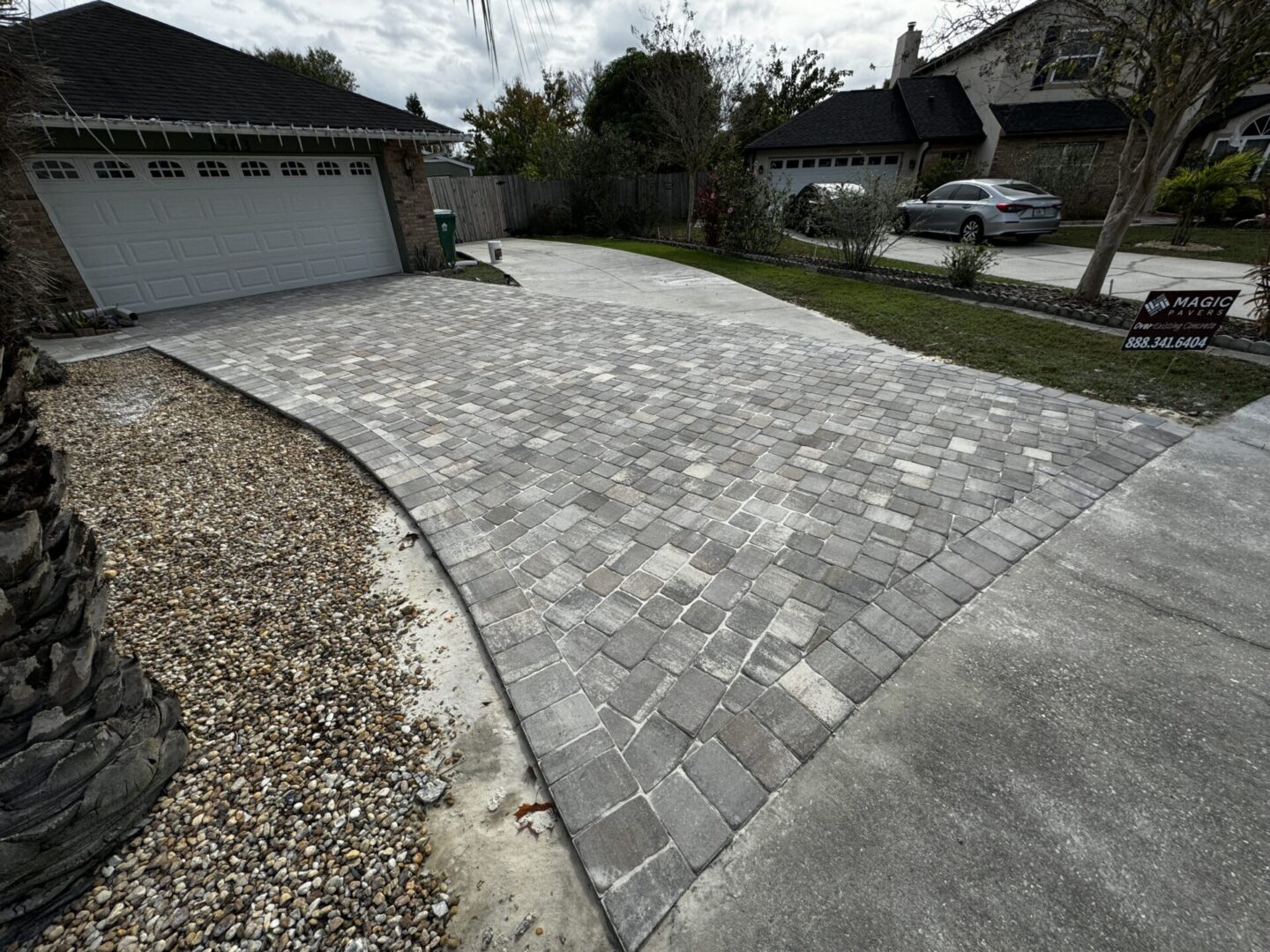 A freshly laid brick driveway leads to a two-car garage. The driveway is bordered by a stone-covered area and a concrete section. A house with a car parked in front is visible in the background.