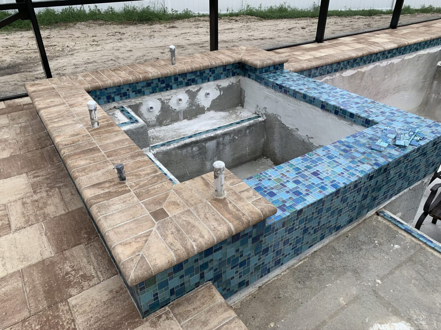 An empty, partially tiled pool under construction, with visible plumbing pipes and unfinished plastering. The pool has blue mosaic tiles and is surrounded by a brick-paved deck.