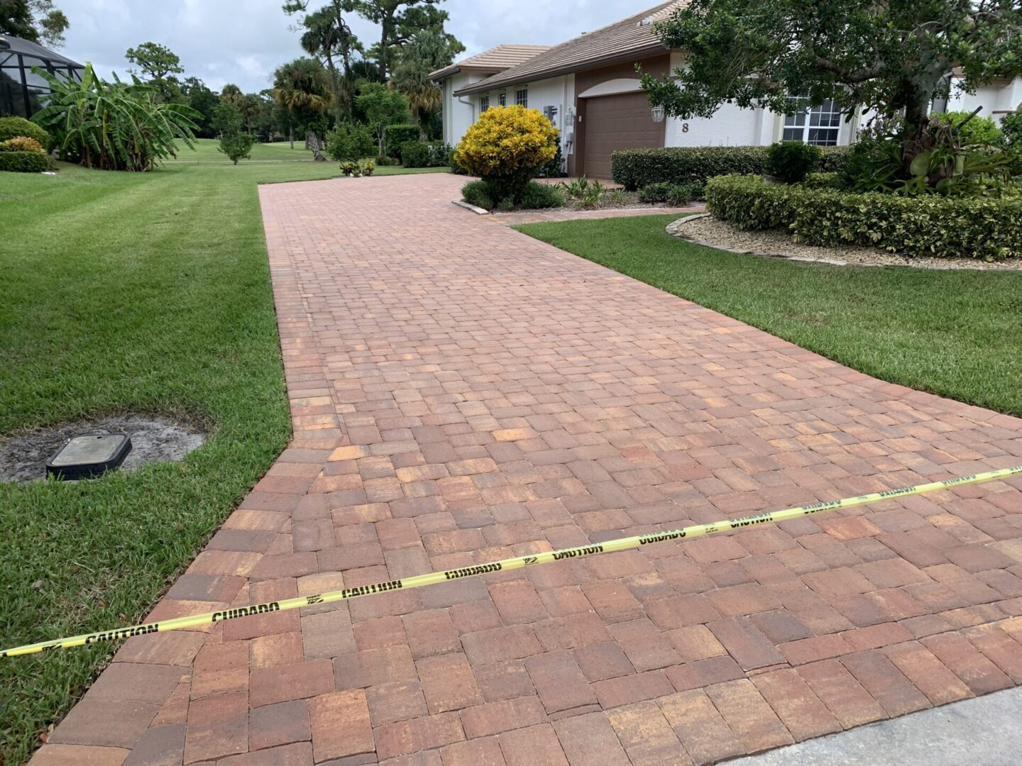 A brick-paved driveway in front of a house is blocked off with yellow caution tape. The surrounding area includes a well-manicured lawn, shrubs, and trees.