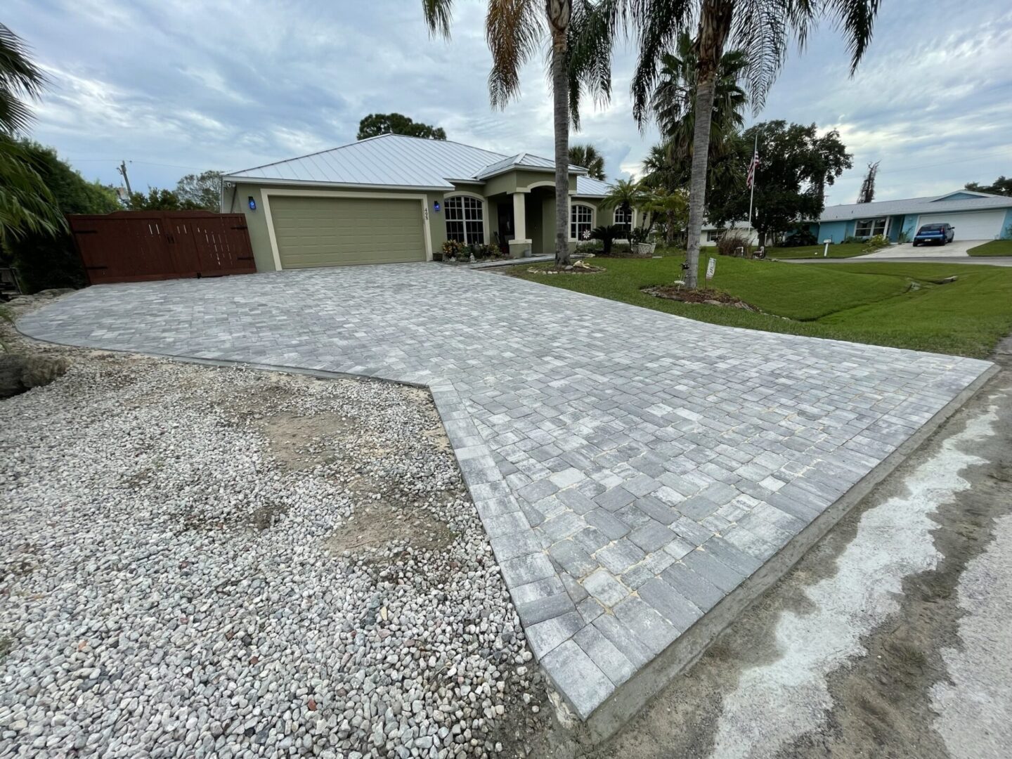 A house with a double garage and a light grey stone paver driveway. There are palm trees and a lawn on the right side. The driveway curves to the left with gravel along the edge.