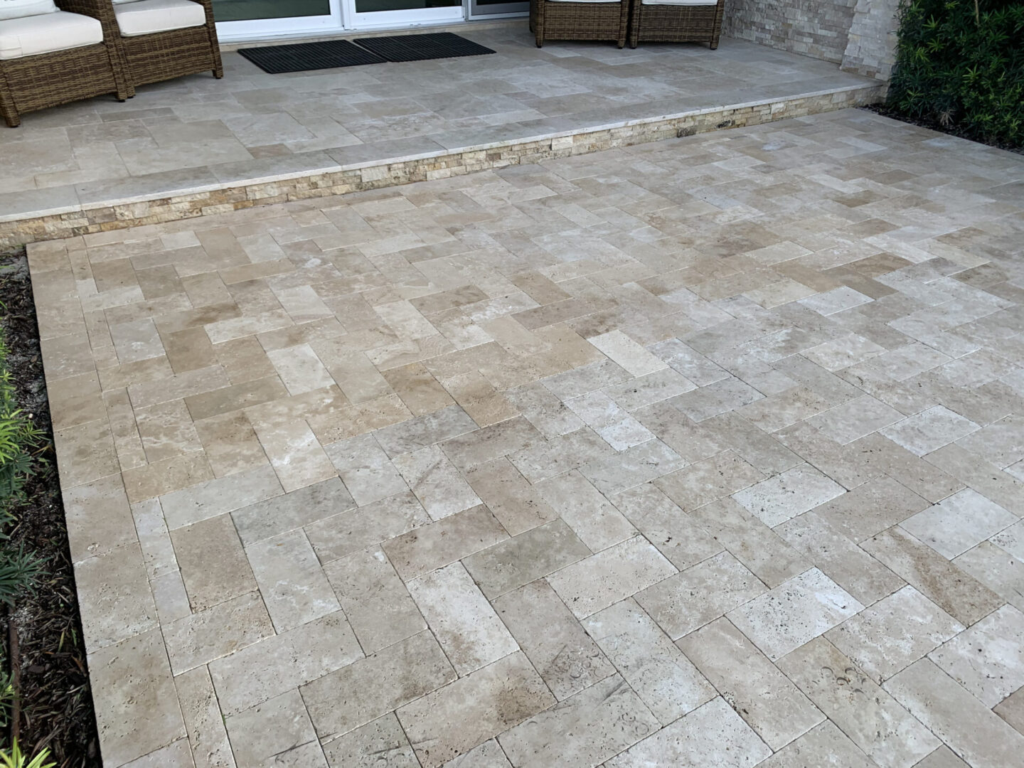 Beige stone tile patio with a step leading up to a seating area with wicker furniture and a sliding glass door.