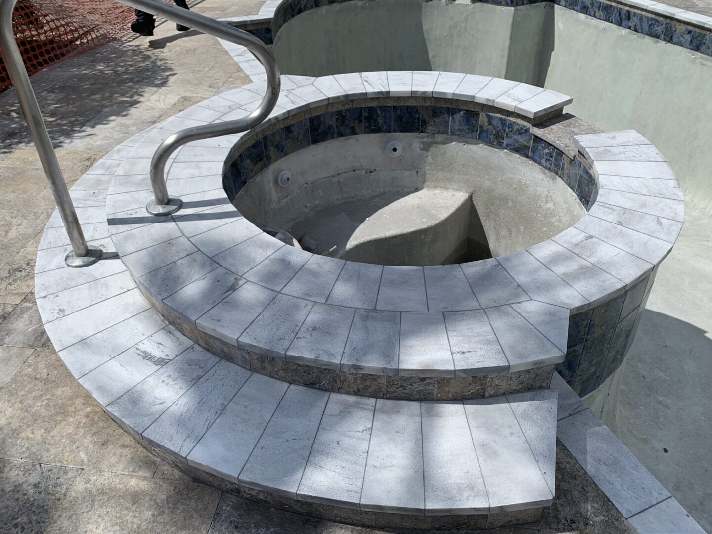 A circular hot tub under construction, featuring a tiled exterior and built-in metal handrails. The interior is not yet finished, showing exposed concrete. The area around is paved.