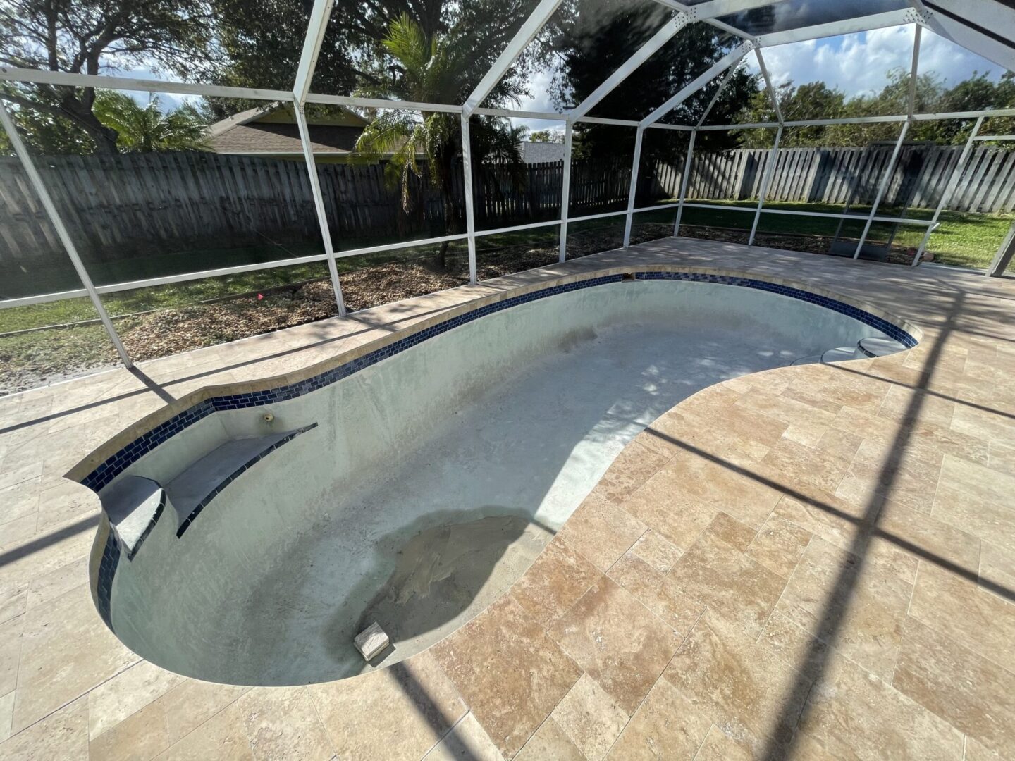 A drained outdoor pool enclosed by a screened fence, with some visible debris at the bottom; surrounded by a stone tile deck. Trees and a wooden privacy fence are in the background.