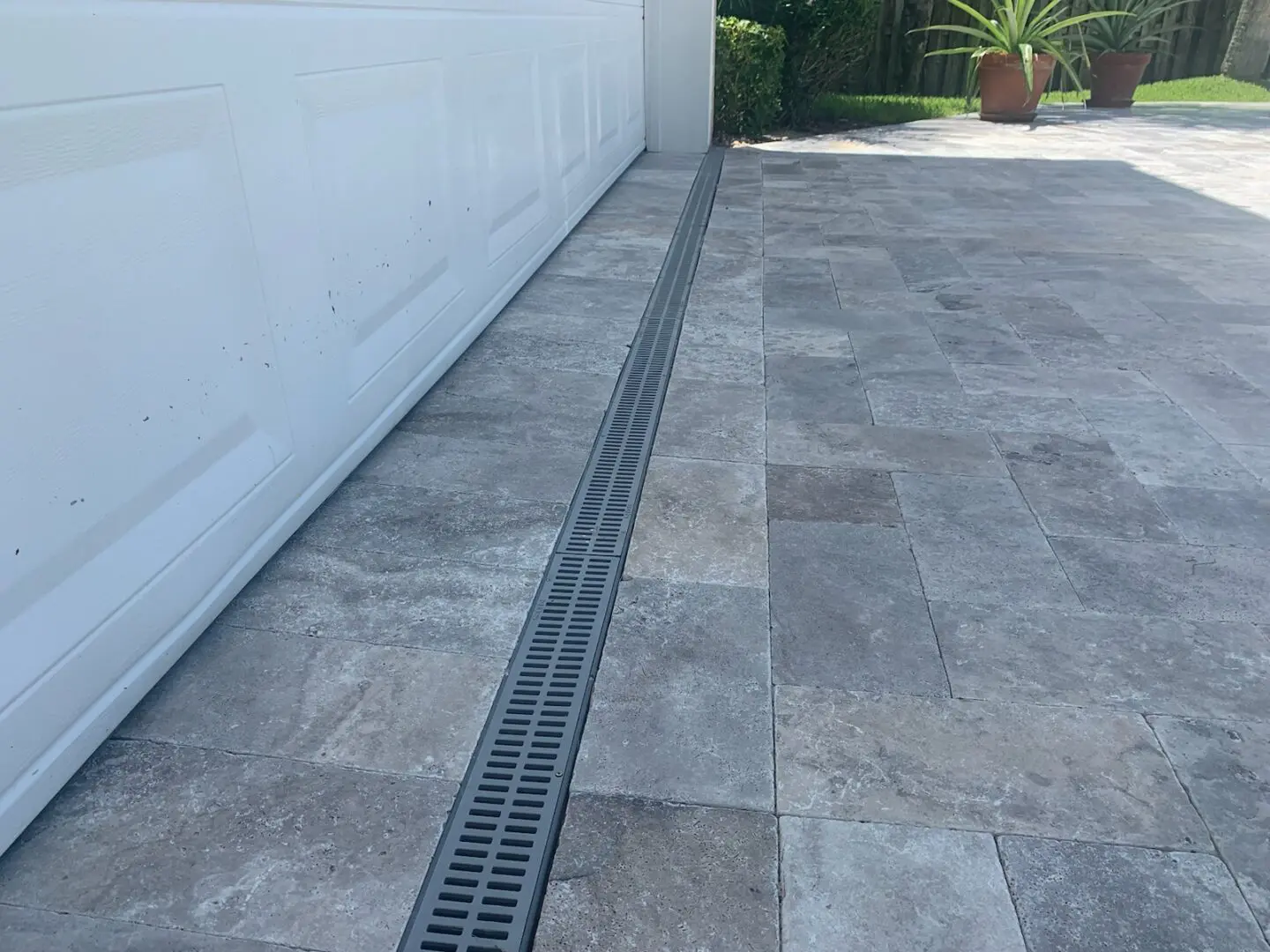 A tiled driveway with a linear drainage grate running parallel to a white garage door. Two potted plants are visible in the background.
