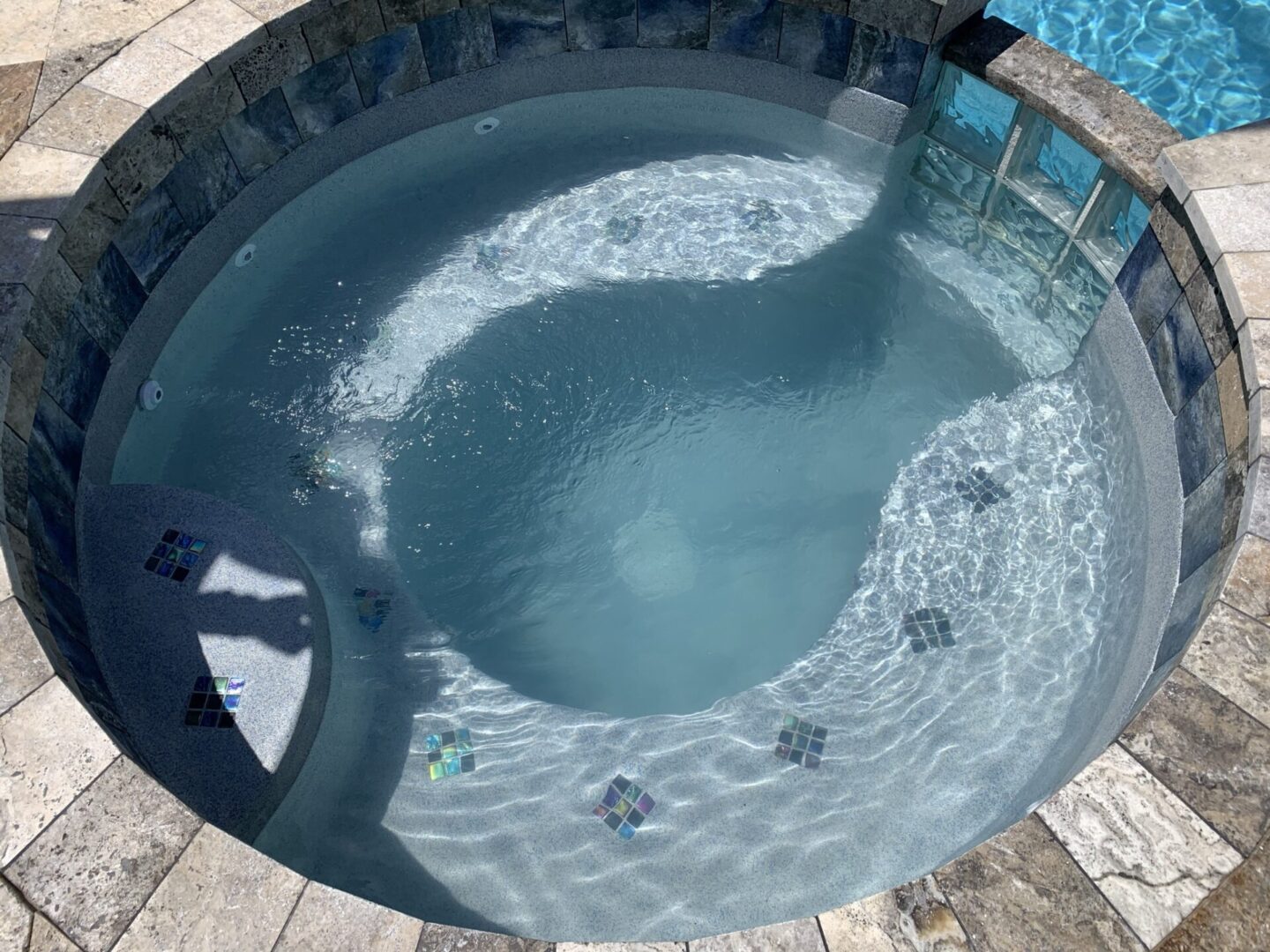 A stone-tiled hot tub filled with water is connected to a swimming pool. Sunlight casts shadows, including one from a nearby person.