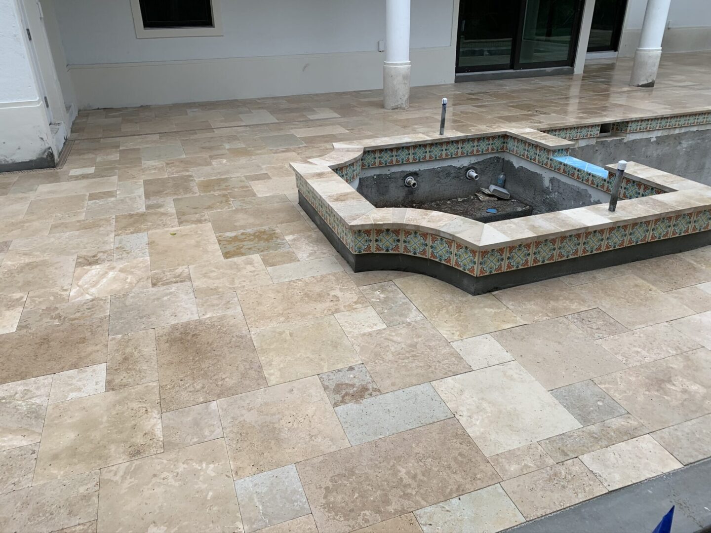 An under-construction tiled patio with an unfinished small rectangular pool featuring decorative tile borders. A building with windows and columns is visible in the background.
