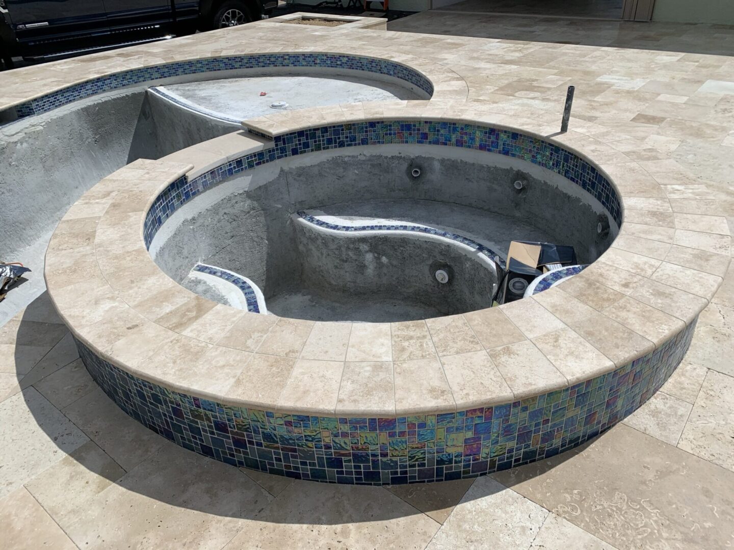 An empty, unfinished circular pool lined with square blue and green mosaic tiles is being constructed on a paved area.