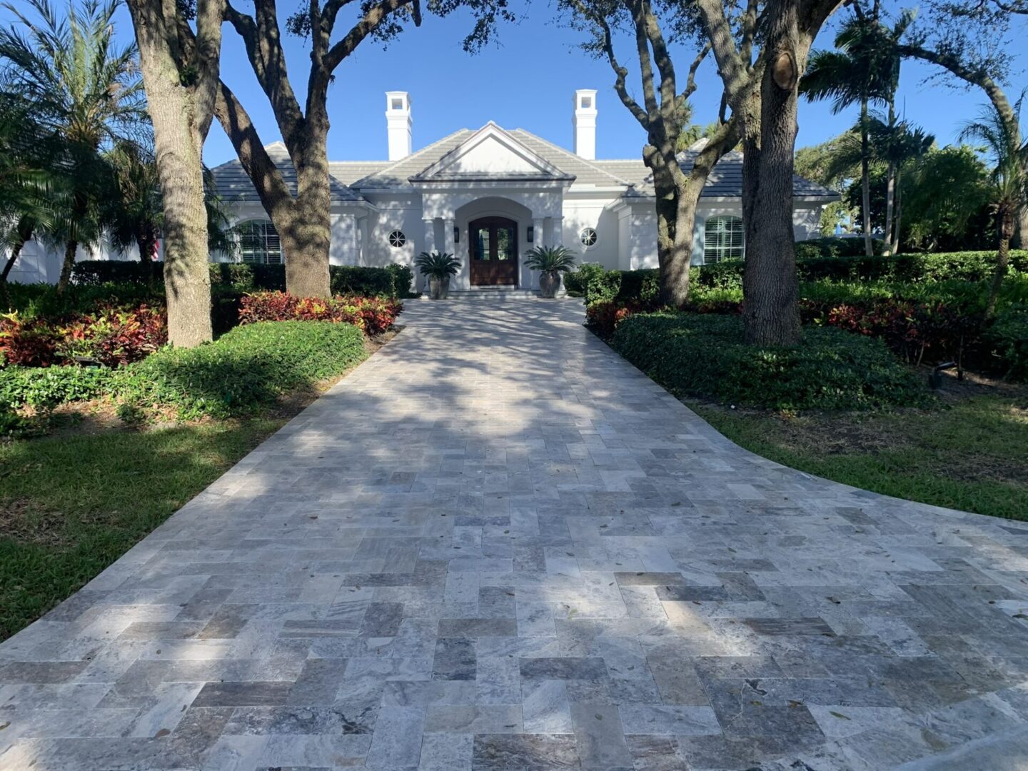A long, stone-paved driveway leads to a grand white house surrounded by trees and lush greenery under a clear blue sky.