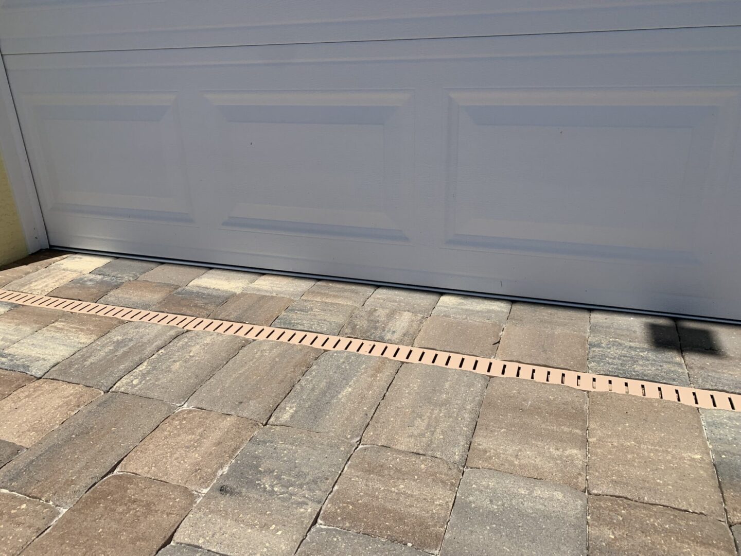 A white garage door with stone brick paving in front and a narrow drainage grate running horizontally across the base.
