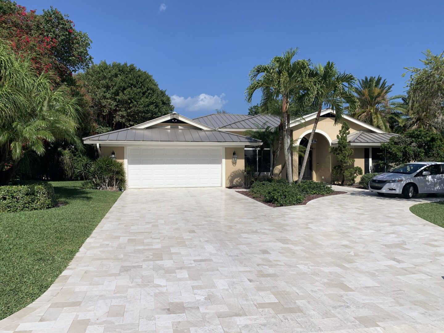 A single-story house with a tiled roof, surrounded by palm trees, featuring a two-car garage and a spacious driveway with a parked car on a sunny day.
