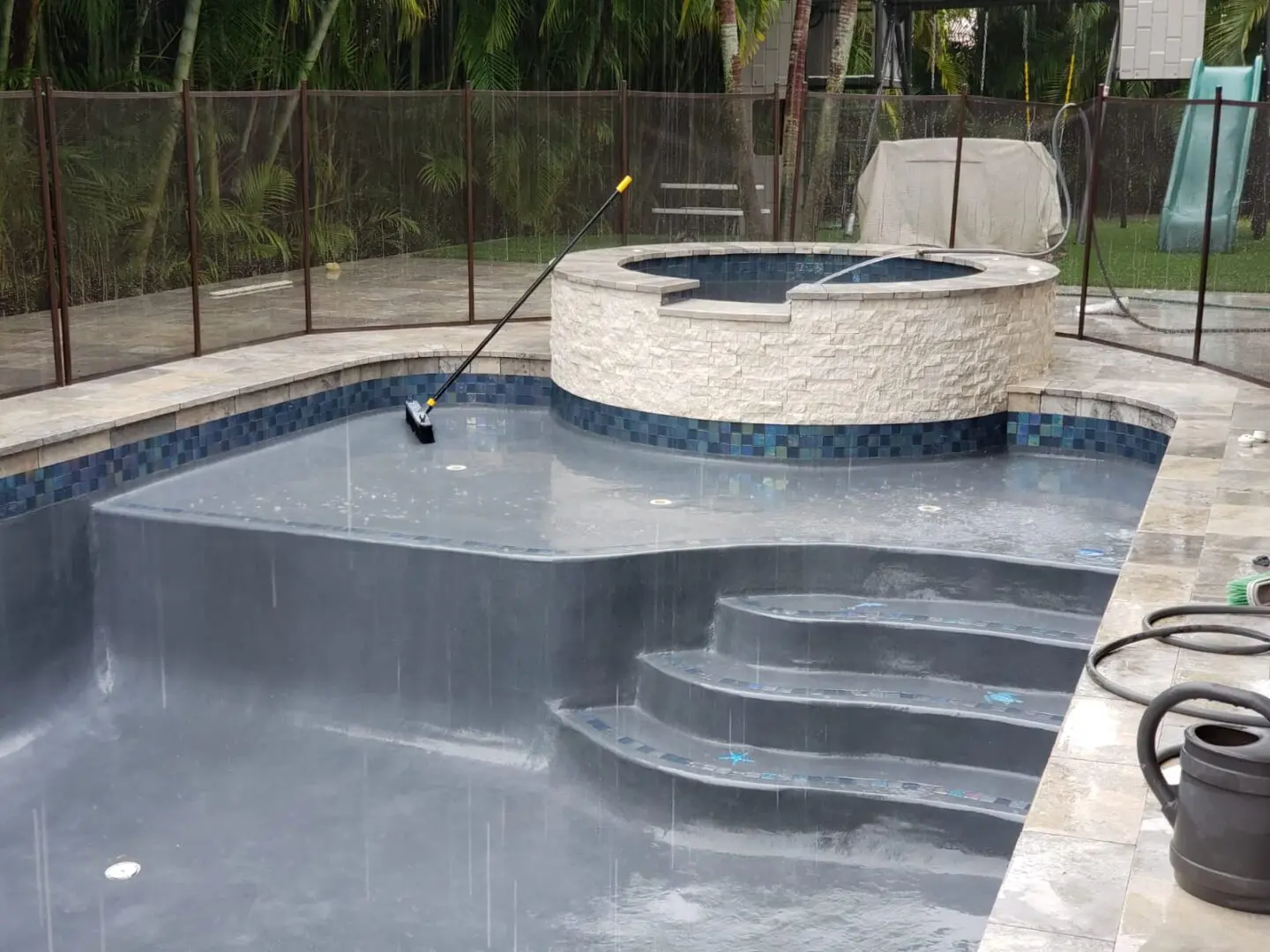 An empty swimming pool and hot tub with a cleaning tool inside, surrounded by a safety fence. The pool and hot tub surfaces are wet, and hoses are lying nearby.