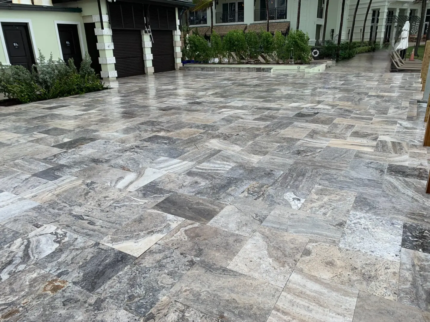 A large driveway paved with interlocking gray stone tiles, leading towards a row of modern houses with garages. The area is clean and wet, indicating recent rainfall or washing.